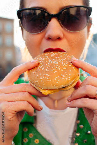 Young pretty girl in sunglasses and green jacket eating big hamburger and looking at camera. Swag brunette woman walking at street  having dinner  eating fast food. Junk food and products.