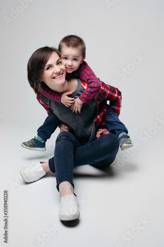 Smiling beautiful brunette mom with little son, sitting on floor at studio, embracing and looking at camera. Parent holding on shoulder her child in checked shirt and jeans. Happy relationship.Gray