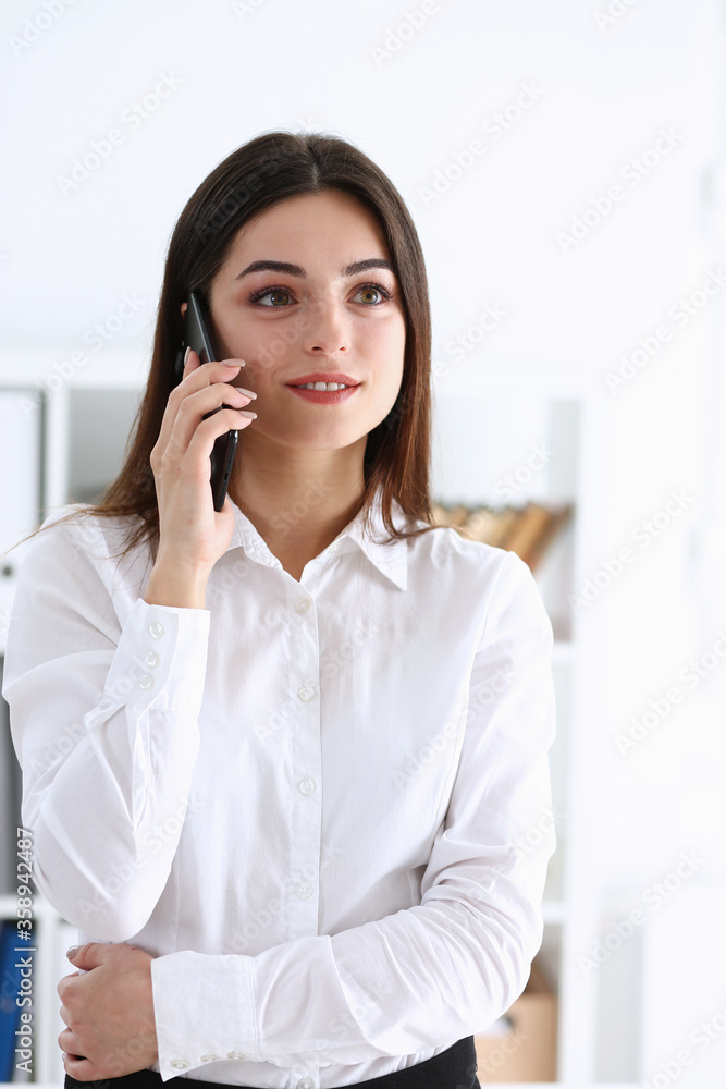 Beautiful businesswoman brunette portrait in an office in a white shirt is holding a phone and talking to a customer remotely looking into the distance listening to what they say and receiving Beautiful businesswoman brunette portrait in an office in a white shirt is holding a phone and talking to a customer remotely looking into the distance listening to what they say and receiving