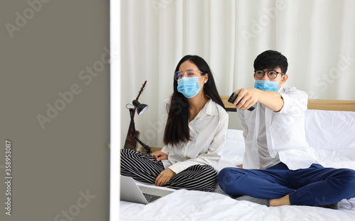 Couple wearing facial mask watching news on television while working at home during quarantine from coronavius or covid-19 pandemic with copy space photo