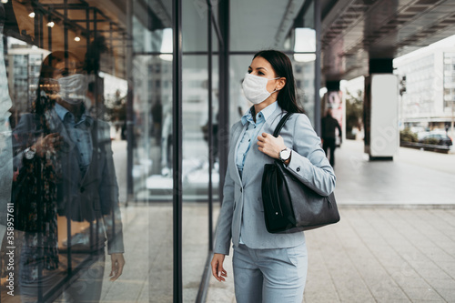 Young and elegant business woman with face protective mask standing on empty street and looking at storefront of closed store. Corona virus or Covid-19 concept.