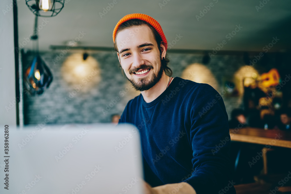 © BullRun - Portrait of cheerful caucasian man in trendy bennie satisfied with online language courses using netbook for studying, happy bearded hipster looking at camera satisfied with wifi for remote work