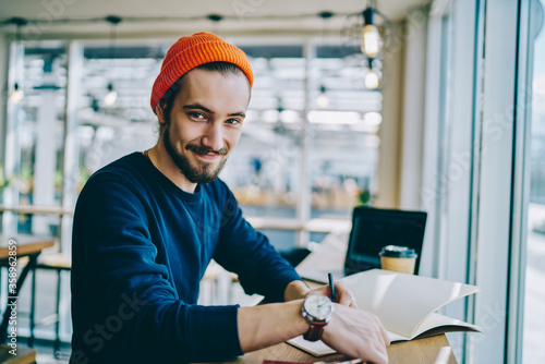 Photos Portrait of cheerful caucasian hipster guy checking time for learning in cafe interior, happy male freelancer looking at camera showing wristwatch using time management for complete remote job