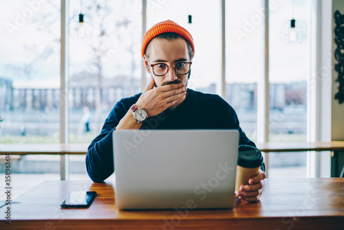 Emotional hipster guy in spectacles read amazing news on laptop computer while working remotely, caucasian man shocked with sales in web store for modern devices and gadgets checking mail on netbook.