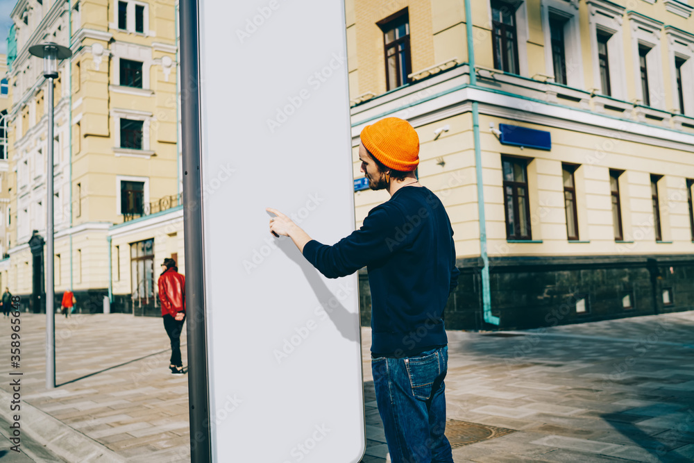 Back view of caucasian male traveler searching information on touristic billboard pointing on white copy space area, hipster guy showing content on blank frame placeholder standing outdoors on street. Back view of caucasian male traveler searching information on touristic billboard pointing on white copy space area, hipster guy showing content on blank frame placeholder standing outdoors on street.