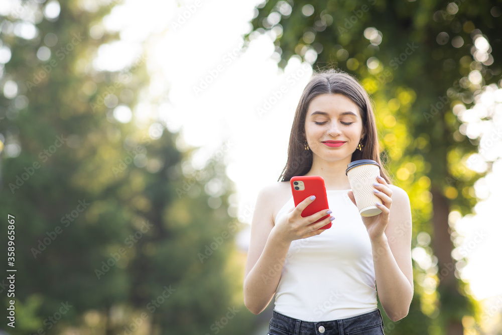 Girl Laughing while looking on her Smartphone. Town Landscape with trees. Cheerful young girl with a paper Cup of coffee in her left hand Girl Laughing while looking on her Smartphone. Town Landscape with trees. Cheerful young girl with a paper Cup of coffee in her left hand