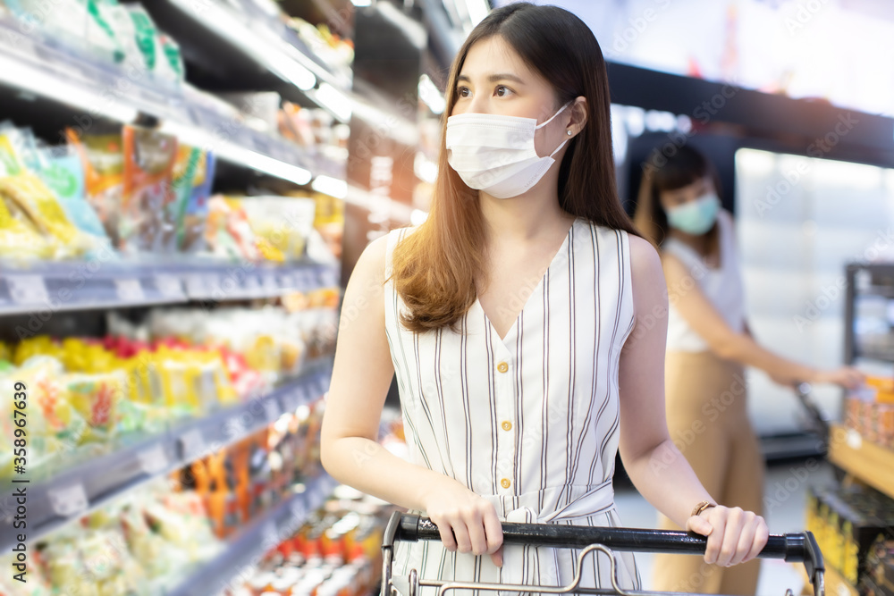 Asian woman wearing face mask push shopping cart in suppermarket . Girl choosing, looking somethings to buy at shelf during coronavirus crisis or covid19 .happy lifestyle or shopaholic concept.