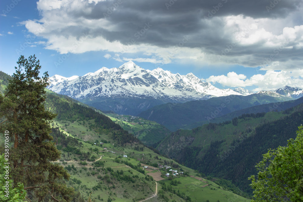 Amazing view of the mountains, wild nature and green valley. Trekking in the mountains. Rural village with farmhouses. View of the countryside. High peaks of the Great Caucasus with snow