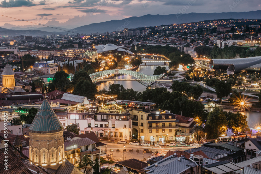 Beautiful panoramic view of evening Tbilisi, capital of Georgia. City colorful lights, river,cathedral,old buildings at a nice sunset mountains in the background.Evening city scene