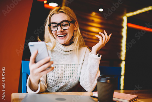 Cheerful hipster girl excited with good news reading mail on smartphone sitting in coffee shop, emotional woman happy about getting discount for internet communication checking news on telephone.