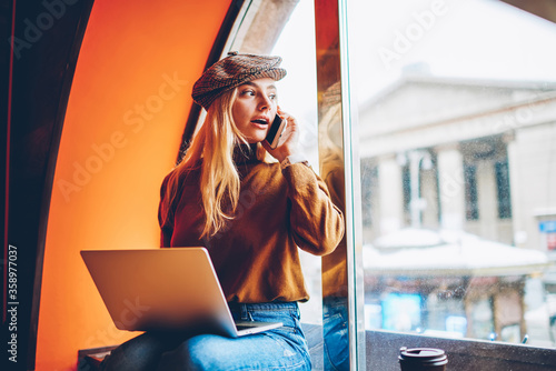 Emotional young woman looking at window surprisely talking on mobile phone during remote job using laptop computer in cafe, hipster girl wondering while calling friend for cellular conversation photo