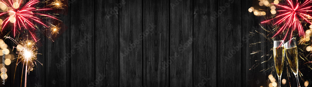 Fototapeta premium Silvester Party Celebration background banner panorama long- Golden red firework and bokeh lights on dark black wooden wall texture and two champagne glasses toasting, with space for text