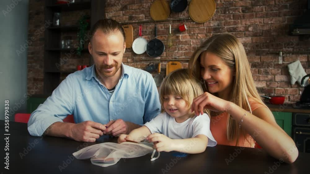 Young spouses and their kid in casual outfit are smiling and playing a game with geometric shapes while sitting at table in kitchen. Close up