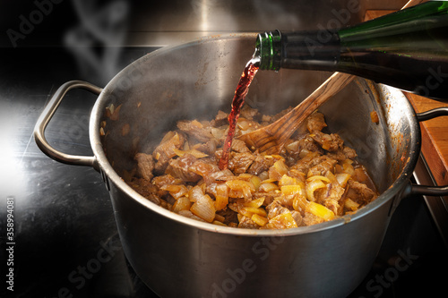 Red wine is poured from a bottle into a pot with roasting goulash to deglaze, cooking at home concept, selected focus