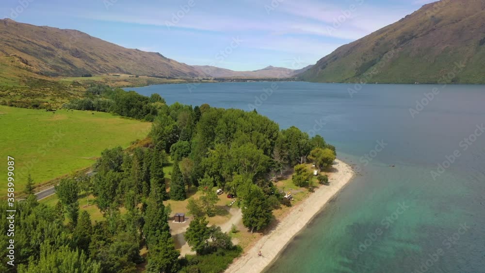 Aerial tilt down shot of vehicles amidst trees at lakeshore, drone flying forward towards mountain range against sky on sunny day - Queenstown, New Zealand Aerial tilt down shot of vehicles amidst trees at lakeshore, drone flying forward towards mountain range against sky on sunny day - Queenstown, New Zealand