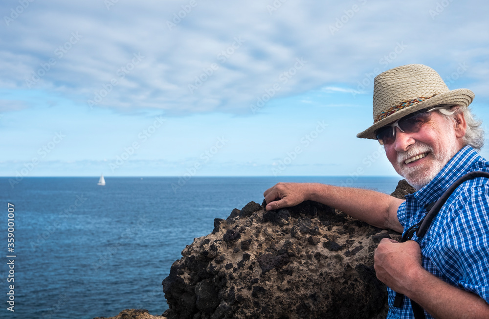 Smiling senior man with straw hat holding a backpack in sea excursion, sailboat and blue sky on background - active retired elderly people and fun concept