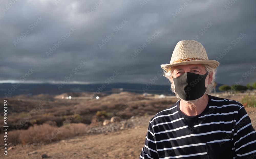 Serious elderly man with straw hat and blue striped shirt in arid landscape wearing a mask because of the Covid-19 coronavirus. Mountains and overcast sky behind him - active retirement concept