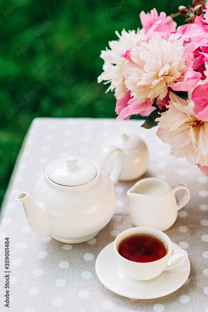 A cozy romantic breakfast in the open air. Peony flower vase, teapot and cup of tea on the table. Congratulations on mother's day or women's day. Soft selective focus. A cozy romantic breakfast in the open air. Peony flower vase, teapot and cup of tea on the table. Congratulations on mother's day or women's day. Soft selective focus.