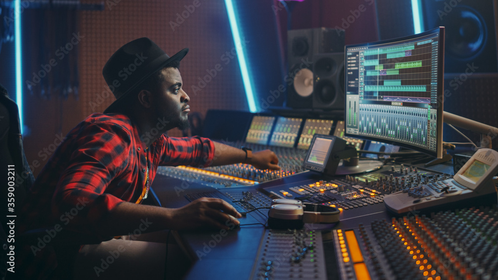 Stylish Audio Engineer Working in Music Recording Studio, Uses Mixing Board and Software to Create Modern Hit Song. Creative Black Artist Musician Working on Desk Control Surface to Produce New Song