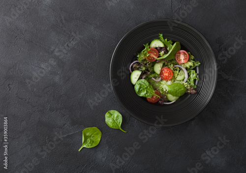 Fresh healthy vegetarian vegetables salad with tomatoes and cucumber, red onion and spinach in black bowl on dark background.