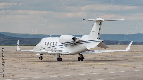 Side view of white small turbofan-powered business jet airplane on the apron of an airport. Cloudy sky. Fast modern aircraft for air transportation. Aviation technology. Travel and business concept.
