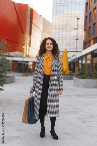 Full length portrait of a beautiful curly young woman with many shopping bags, over buildings background.