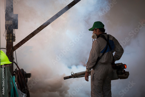 Asian healthcare worker in protective clothing using fogging machine spraying chemical to eliminate mosquitoes and prevent dengue fever inside of warehouse area
