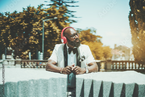 Handsome mature bearded bald African man in glasses and an elegant white suit is enjoying sunny summer weather, standing outdoors, and listening to music in his reddish headphones