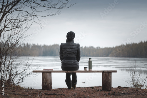 A young woman sits on a bench on the shore of a frozen lake. Thermos and a book are nearby. Woman in focus blurred background.