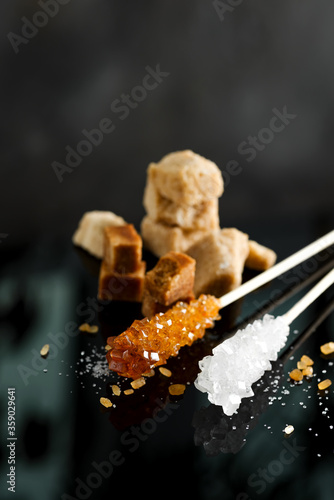 Various types of sugar brown cane cubes and crystal sugar sticks on a black background with reflection, copy space.