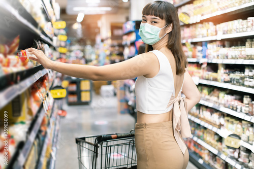 Asian woman wearing face mask push shopping cart in suppermarket . Girl choosing, looking somethings to buy at shelf during coronavirus crisis or covid19 .happy lifestyle or shopaholic concept.