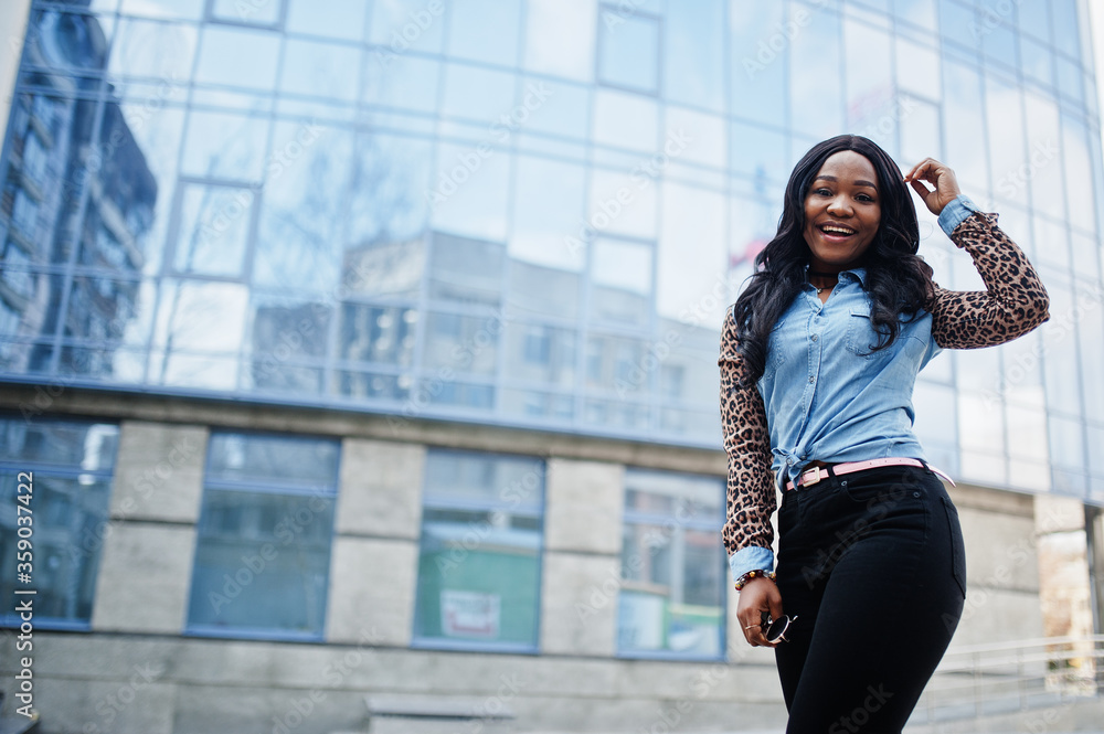 Hipster african american girl wearing jeans shirt with leopard sleeves posing at street against modern office building with blue windows. Hipster african american girl wearing jeans shirt with leopard sleeves posing at street against modern office building with blue windows.