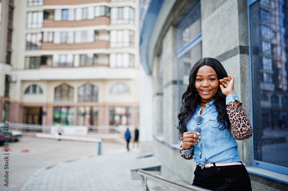 Hipster african american girl wearing jeans shirt with leopard sleeves posing at street against modern office building with blue windows.