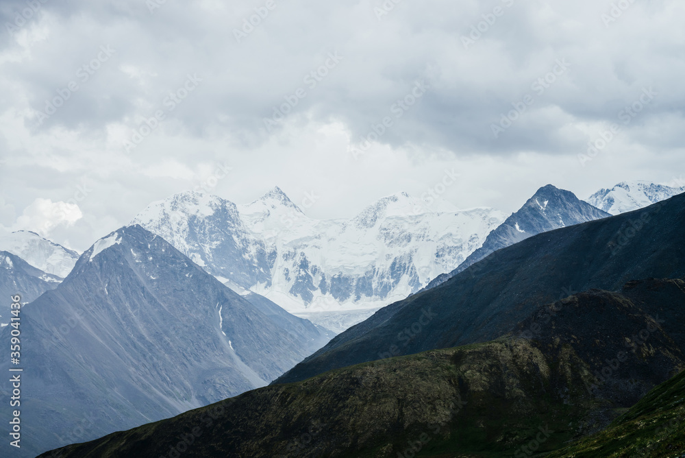 Fototapeta premium Wonderful view to huge glaciers on great snowy mountain range behind big rocks under gloomy cloudy sky. Pointy tops of giant rocky mountains. Awesome beauty of highlands. Atmospheric alpine landscape.