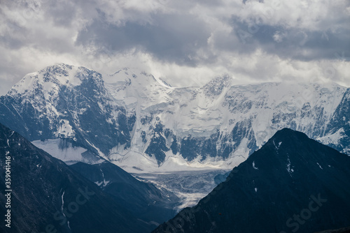 Awesome landscape with huge glacial mountains in bad cloudy weather. Low stormy clouds touch top of snowy mountain with glaciers. Storm is coming due to mountains. Gloomy overcast atmospheric scenery.