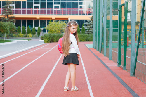 little sad girl in school uniform with a backpack outside near the school