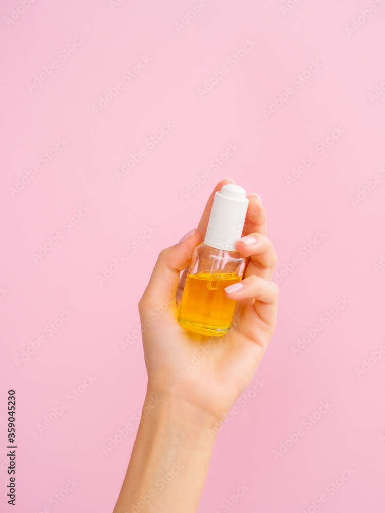 Close up cosmetic oil products with copy space for your logo. Feminine hands hold clear dropper bottle with yellow oil on a pink background Close up cosmetic oil products with copy space for your logo. Feminine hands hold clear dropper bottle with yellow oil on a pink background
