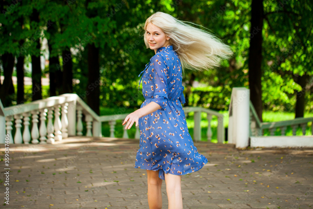 Portrait of a young blonde woman in blue dress walking in summer parkpark