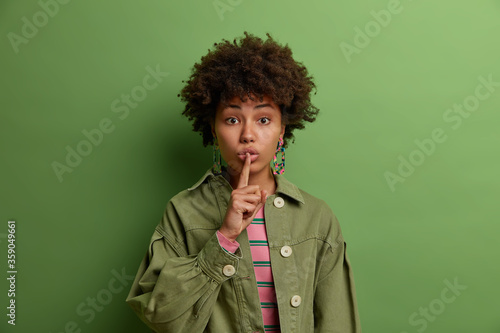 Mysterious beautiful woman with Afro hair presses index finger to folded lips makes hush gesture and asks to be quiet asks politely to keep silence prohibits talking during meeting wears green clothes