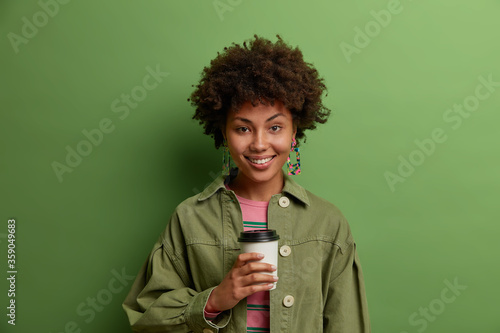 Happy brunette ethnic woman holds disposable cup of coffee, enjoys break, smiles positively, tastes aromatic beverage, wears green jacket, stands indoor. Girlfriend poses with caffeine drink
