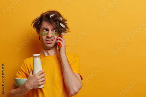 Displeased man has bad mood in morning, going to have breakfast, talks via smartphone, wears collagen pads under eyes, busy and awakes late, isolated on yellow background. Morning routine concept © Wayhome Studio