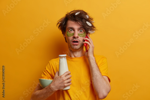 Shocked hipster guy finds out bad news in morning, has phone conversation with friend, holds breakfast ingredients for energy charge, cares about skin, wears patches under eyes, poses indoor. © wayhome.studio 