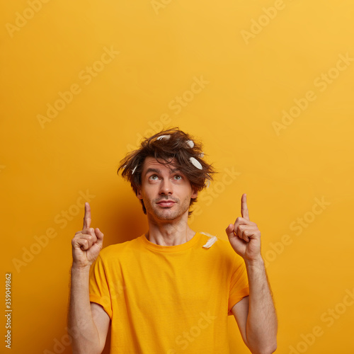 Curious man with messy hair, feathers, indicates both index fingers upwards, shows empty space for your advertisement, recommends nice offer, wears casual yellow t shirt, stands indoor. Promo here