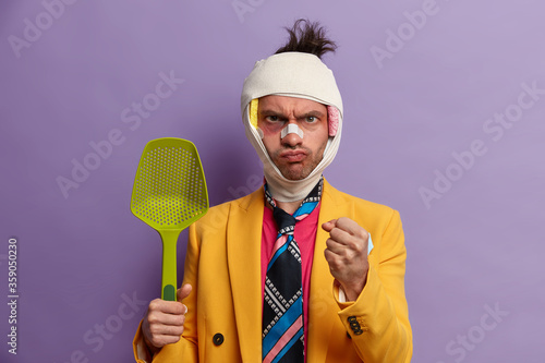 Strict serious man clenches fist, ready for fight and defence himself, being abused by someone, beaten and has shiners, head conscussion, wears colorful tie and yellow jacket, holds kitchenware photo