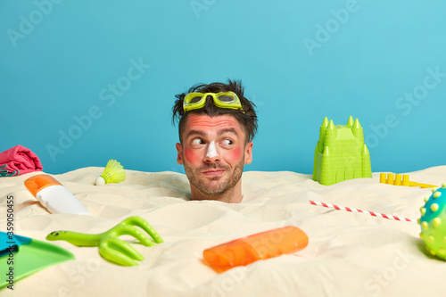 Принти на полотні Funny positive man has head stuck in sand, wears goggles, sunburnt red skin, rel