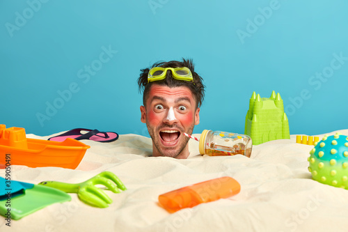 Happy crazy man sunbathes in sun, has danger of burns, buried in sand, looks positively at camera, enjoys summer recreation, ideal sunny hot day at beach, has active rest, uses protective sunscreen