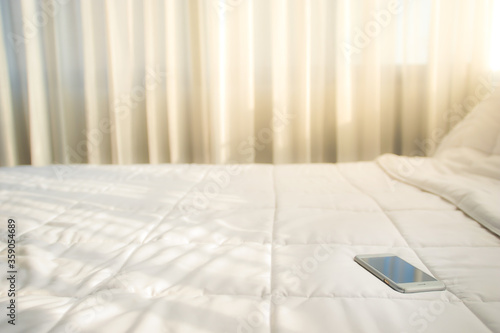 The smartphone is placed on a bed with a white sheet. In a room with white curtains and sunlight shining through the curtains photo