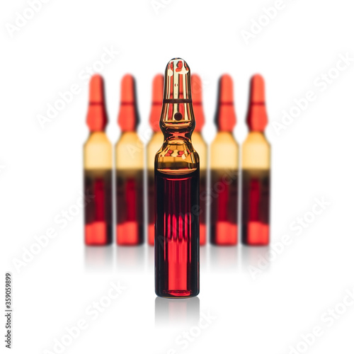 An orange glass medical bottle in front of blurred medical ampoules in a row with red liquid. Isolated on white