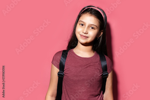 Studio portrait of cheerful child girl wearing headband and backpack on background of pastel pink color.