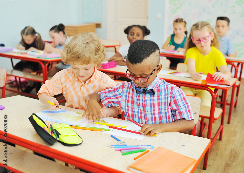 Fototapeta premium two small children of elementary school students are preparing for a lesson sitting at a Desk with colored pencils in their hands against the background of a group of children in the classroom.